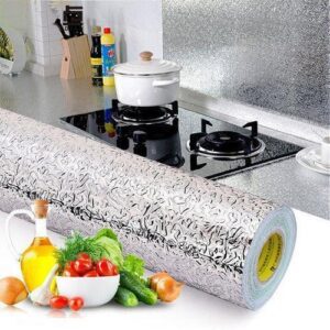 Se coloca un rollo de papel de aluminio decorativo con un reverso adhesivo sobre una encimera de cocina con verduras frescas, aceite y condimentos cerca, lo que sugiere una mejora en la decoración de la cocina o una sesión de cocina a punto de comenzar.