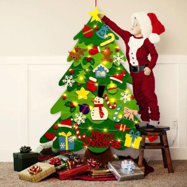 Un niño pequeño vestido con un traje festivo de Papá Noel extendiendo la mano para colocar la estrella encima de un colorido árbol de Navidad de fieltro, rodeado de regalos envueltos.