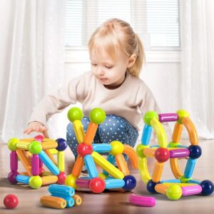 A young child with blonde hair wearing a beige sweater and blue polka dot pants is sitting on the floor, playing with colorful magnetic building toys. The room has large windows with blinds and white curtains, letting in plenty of light.