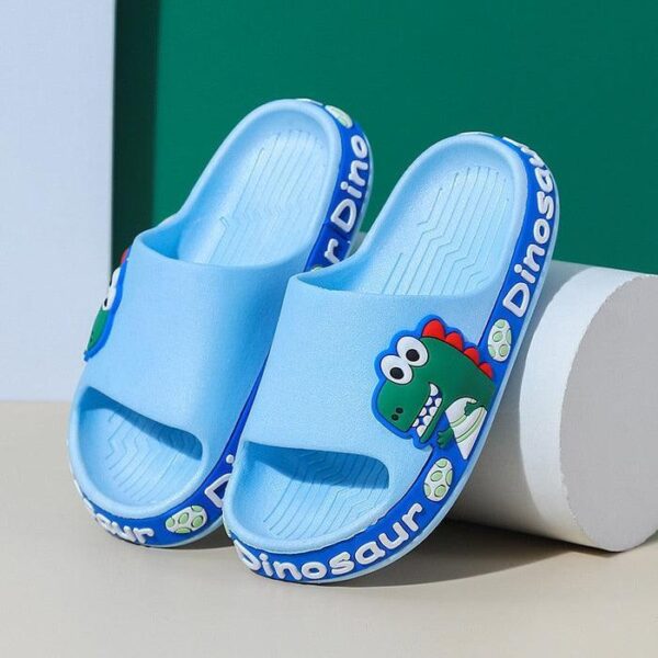 Un par de pantuflas infantiles azules con un lindo diseño de dinosaurio, exhibidas frente a un fondo verde y blanco.