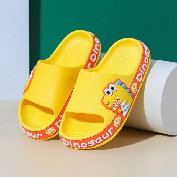 Pantuflas infantiles de color amarillo brillante con un divertido diseño de dinosaurio, sobre un fondo verde y blanco.