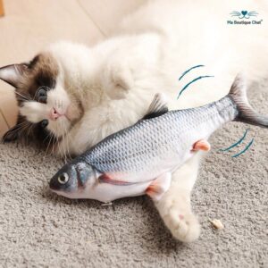 A white and brown cat with blue eyes is lying on a grey carpet, playfully interacting with a realistic-looking toy fish. The fish appears to be designed to move, resembling a wriggling motion. The background shows a wooden floor.