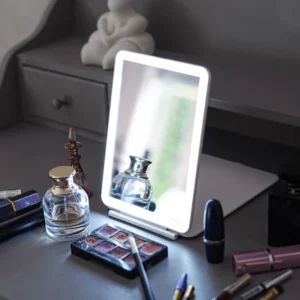 A lighted vanity mirror on a gray table surrounded by various makeup items, including eyeshadow palettes, lipstick tubes, and perfume bottles. An abstract sculpture is visible in the background on a drawer unit.