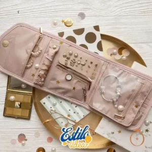 A pink travel jewelry box - EstiloBag, with various compartments is laid open on a decorative surface. It contains earrings, bracelets, and necklaces. Surrounding it are metallic plates, polka dot paper, and scattered sequins. The words "Travel jewelry box - EstiloBag" appear on the bottom.