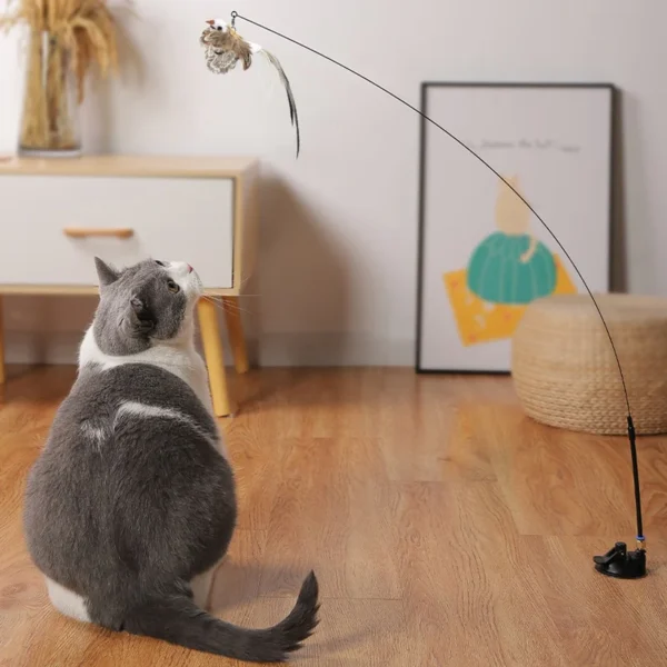 Un chat gris et blanc est assis sur un plancher en bois, regardant attentivement une souris suspendue à un jouet pour chat en forme de canne à pêche. Le jouet est soutenu par un support. En arrière-plan, il y a un caisson à tiroirs blanc et une image encadrée d'une pastèque sur le mur.